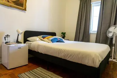 a bedroom with a bed and a lamp and a window at Aconchego de quartos no centro de Faro in Faro