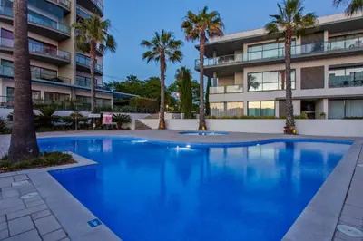 a swimming pool with palm trees in front of a building at Areias Branca in Quarteira