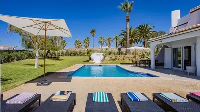 a swimming pool with chairs and an umbrella next to a house at Greice Homes - Villa Carob Tree in Almancil