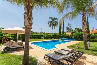 a swimming pool with two chairs and a umbrella and two palm trees at Fonte Santa Villa in Quarteira