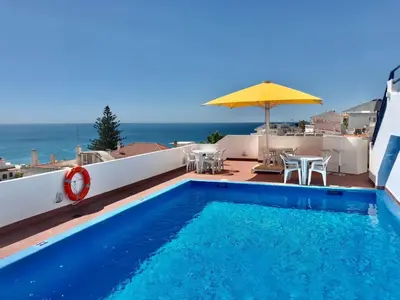 a blue swimming pool with an umbrella and the ocean at Hotel da Gale in Albufeira