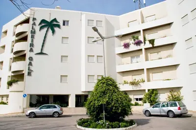 two cars parked in front of a building with a palm tree at O Apartamento A Ramos Palmeira Galé Está inserido num Condomínio com PISCINA, Fica apenas a 250 Metros da Praia da Galé - Apartamento com Ar Condicionado na Sala, Smart TV com Prime Vídeo e Cozinha Completa in Albufeira