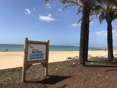 a sign on a beach with palm trees and the ocean at Apartamento frente ao mar com piscina de água salgada in Quarteira