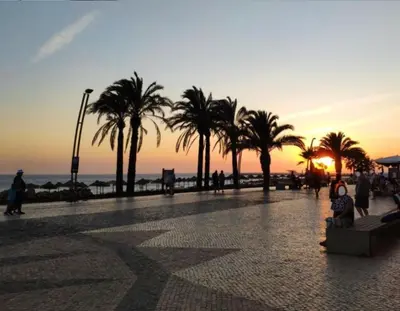 people walking on a beach with palm trees at sunset at Seaside Serenity: 1BR with Breathtaking Ocean View in Quarteira
